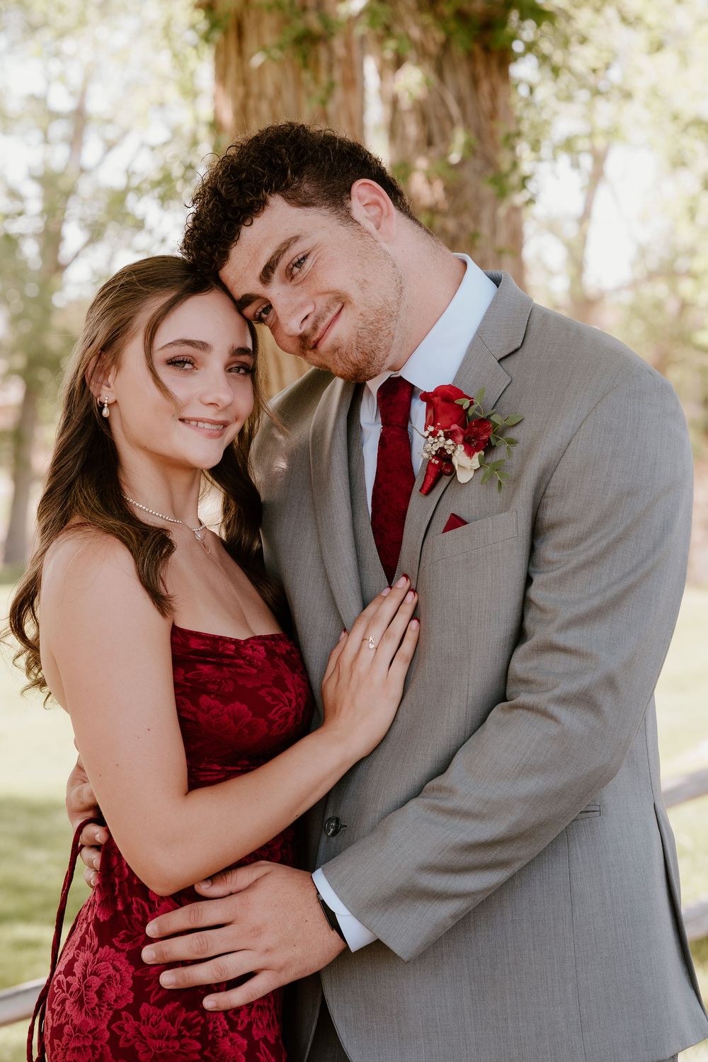 A couple in formal attire embraces outdoors, man in gray suit with red tie and boutonniere, woman in red lace dress.
