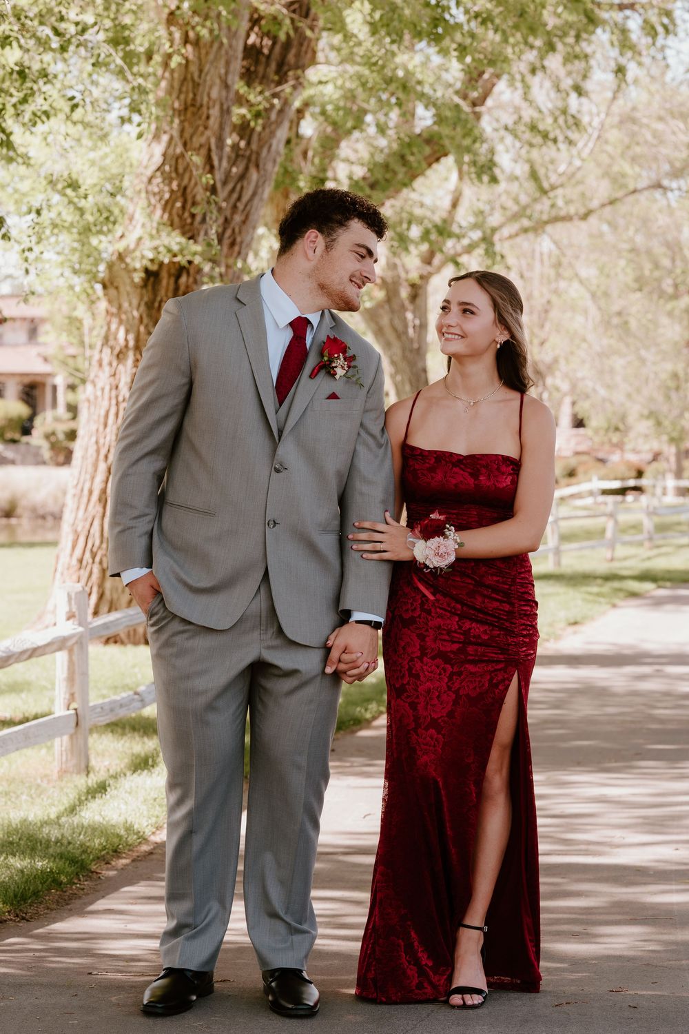 A couple in formal prom attire walk hand-in-hand, man in gray suit, woman in red gown, outdoors.