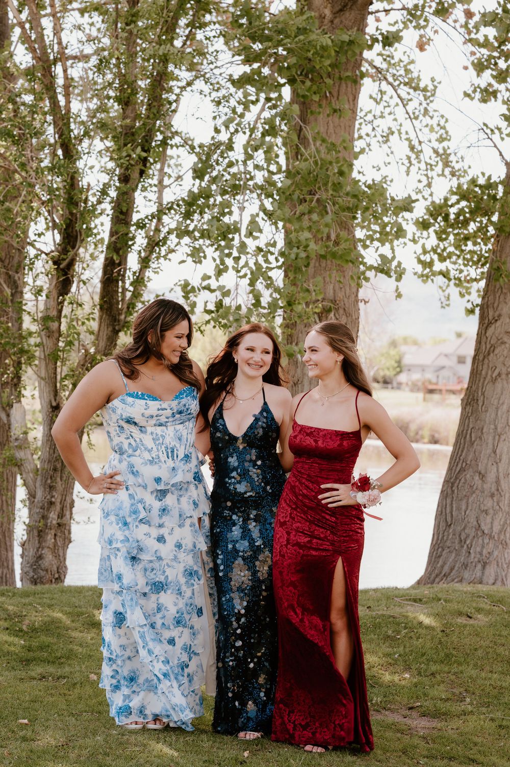 Three teenage girls in elegant prom dresses posing outdoors among trees, wearing blue floral, navy sequin, and red velvet gowns.