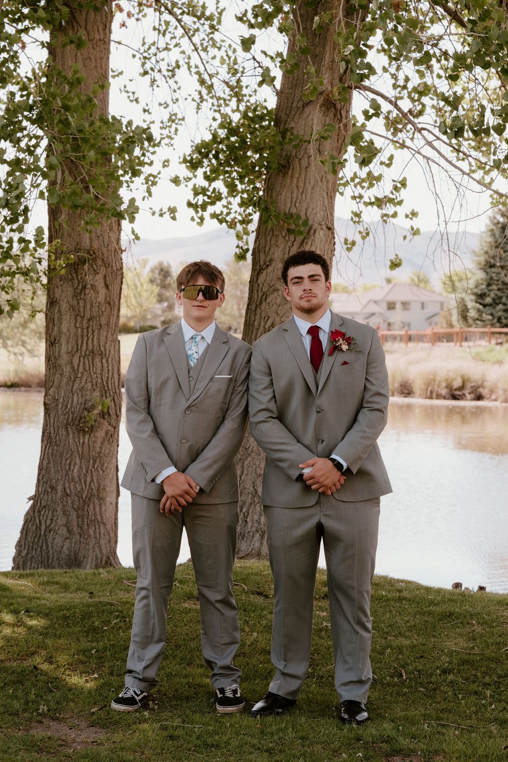 Two young men in matching gray suits with red boutonnieres standing by trees near a lake.