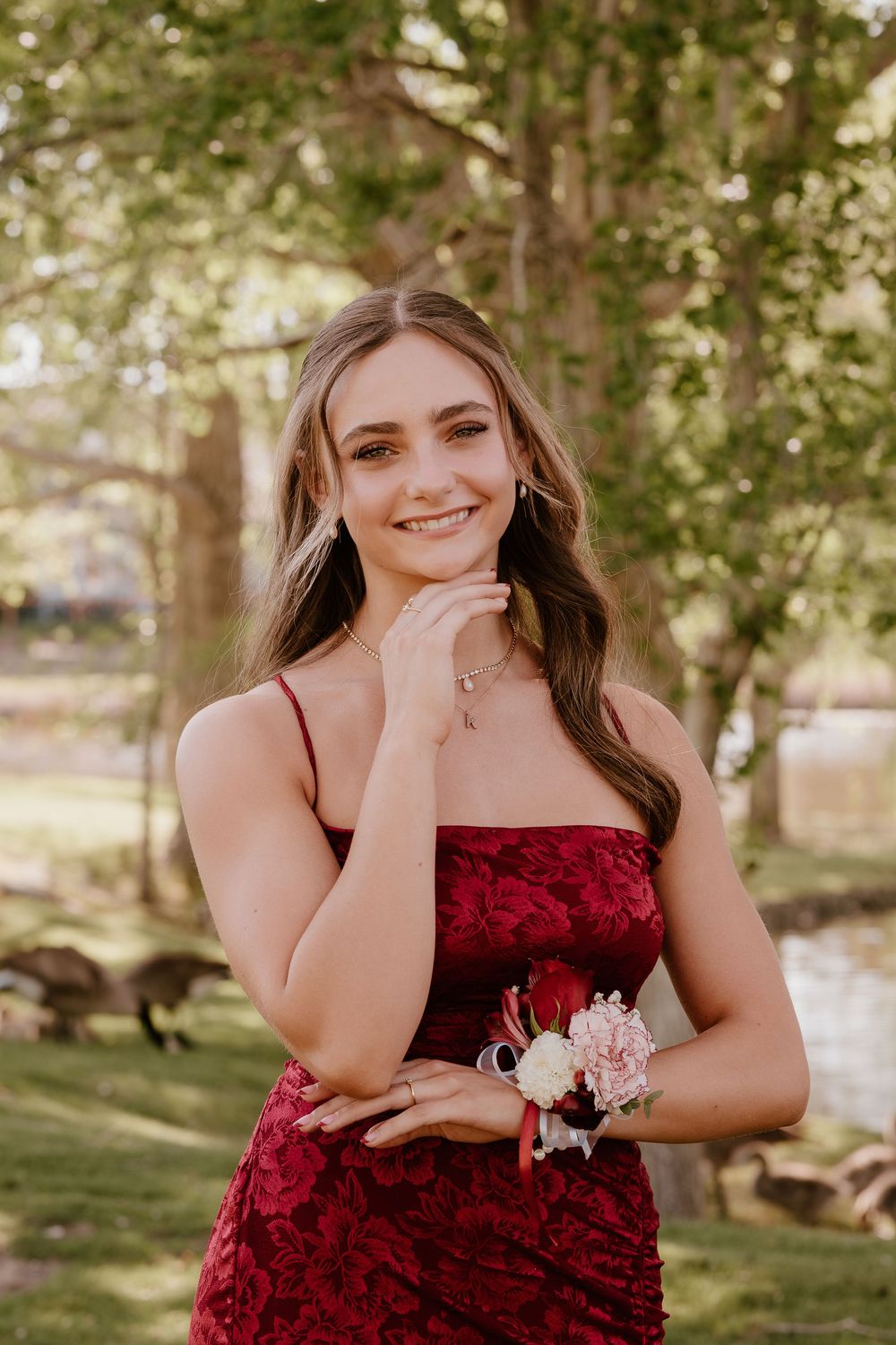 A smiling young woman in a red lace gown poses outdoors wearing a floral wrist corsage.
