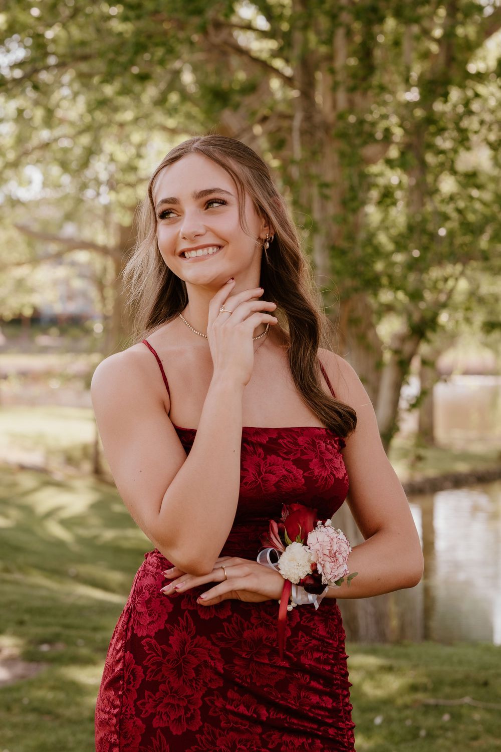 Young woman in red lace gown with floral wrist corsage smiling outdoors near trees at prom.
