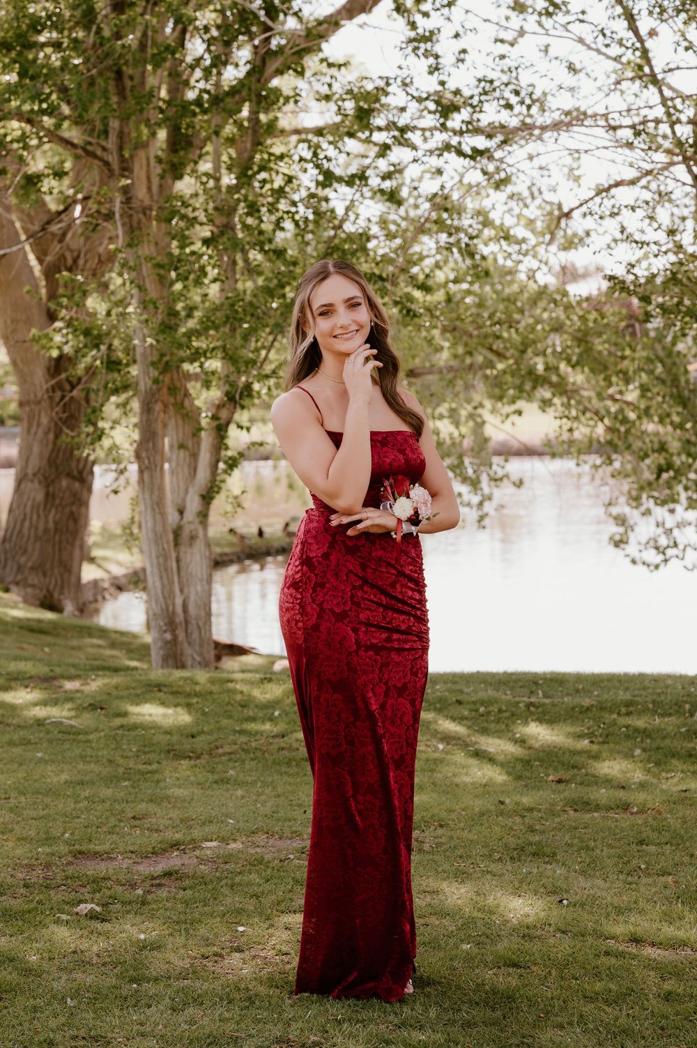 Young woman in a red sequin gown holding a corsage, posing outdoors near a river with lush green trees.