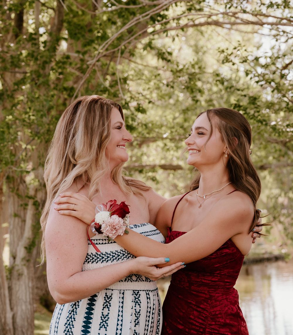 Two women in formal dresses smile at each other outdoors, one wearing a floral corsage, surrounded by trees.