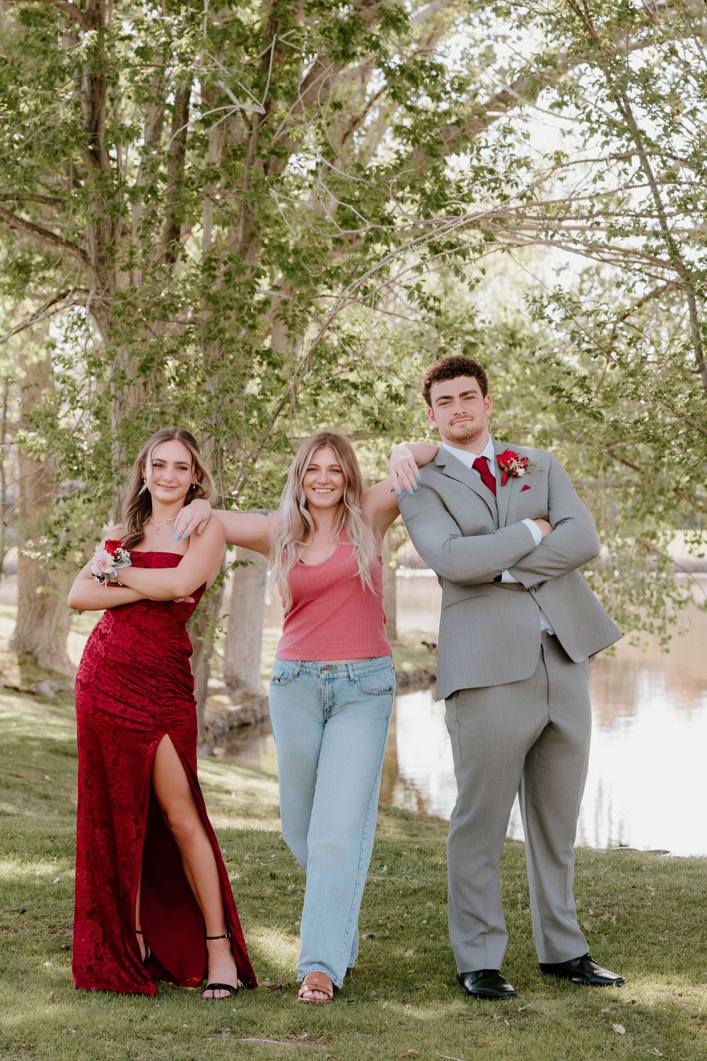Two prom-dressed teens and a friend pose outdoors near a pond, wearing red sequin gown, gray suit, and casual attire.