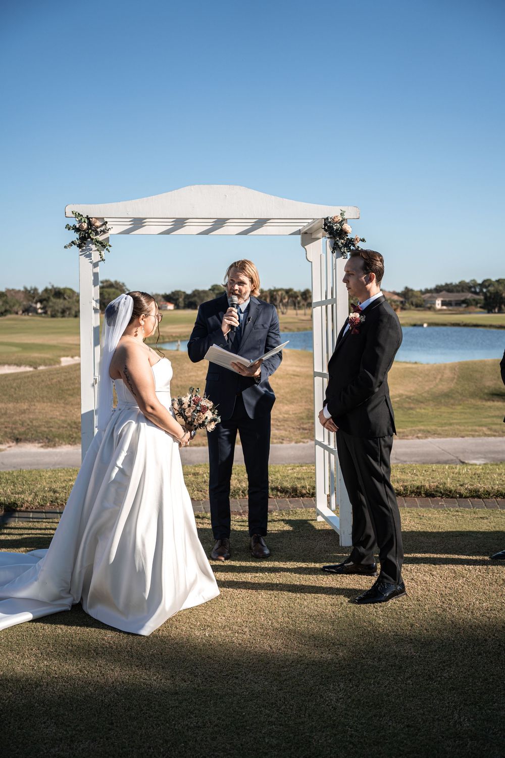 Bride and groom exchange vows under a white arch on a sunny golf course with an officiant holding a microphone.