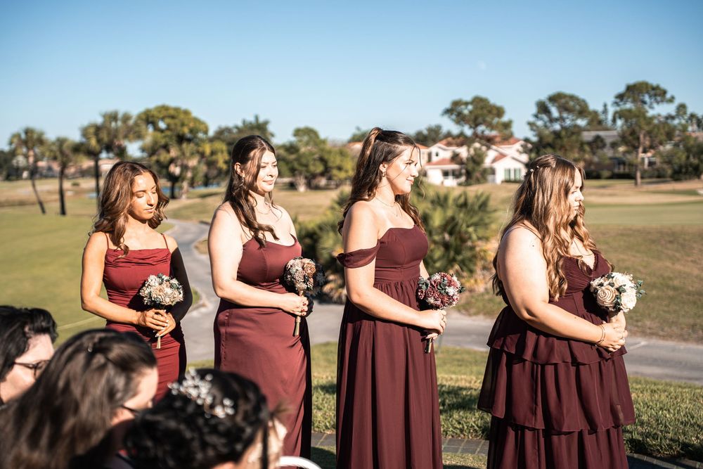 Four bridesmaids in burgundy dresses holding bouquets stand outdoors at a wedding ceremony on a sunny day.