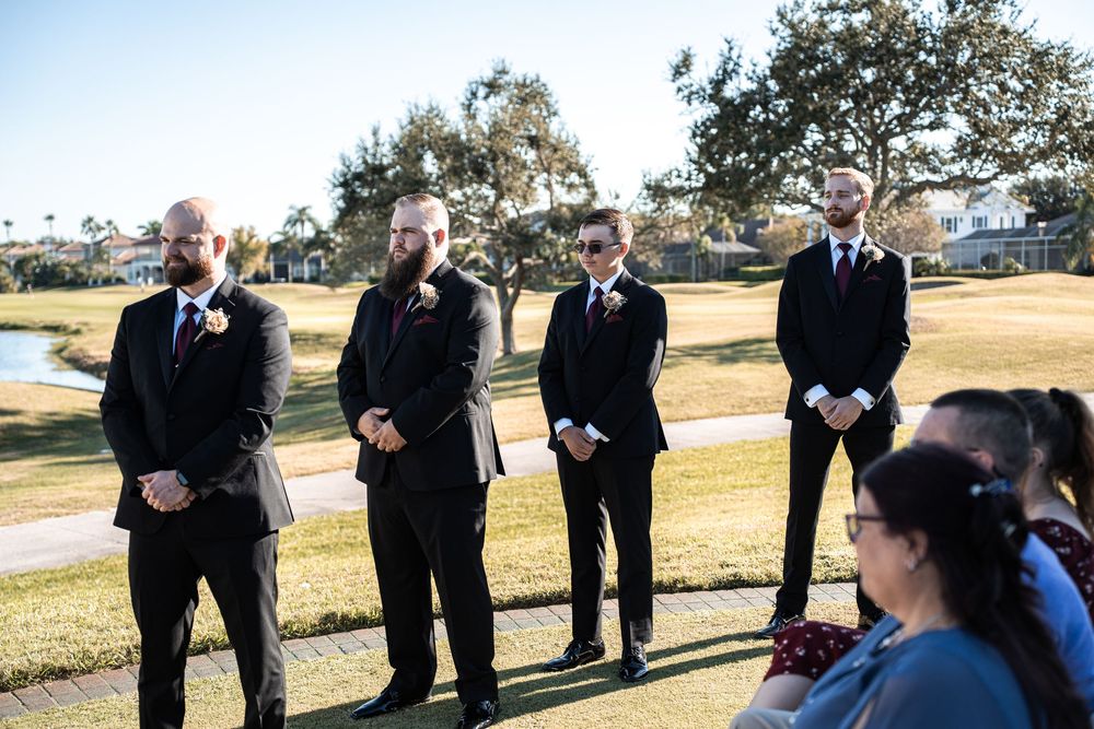 Four groomsmen in black suits with red boutonnieres stand outdoors at a golf course wedding ceremony.