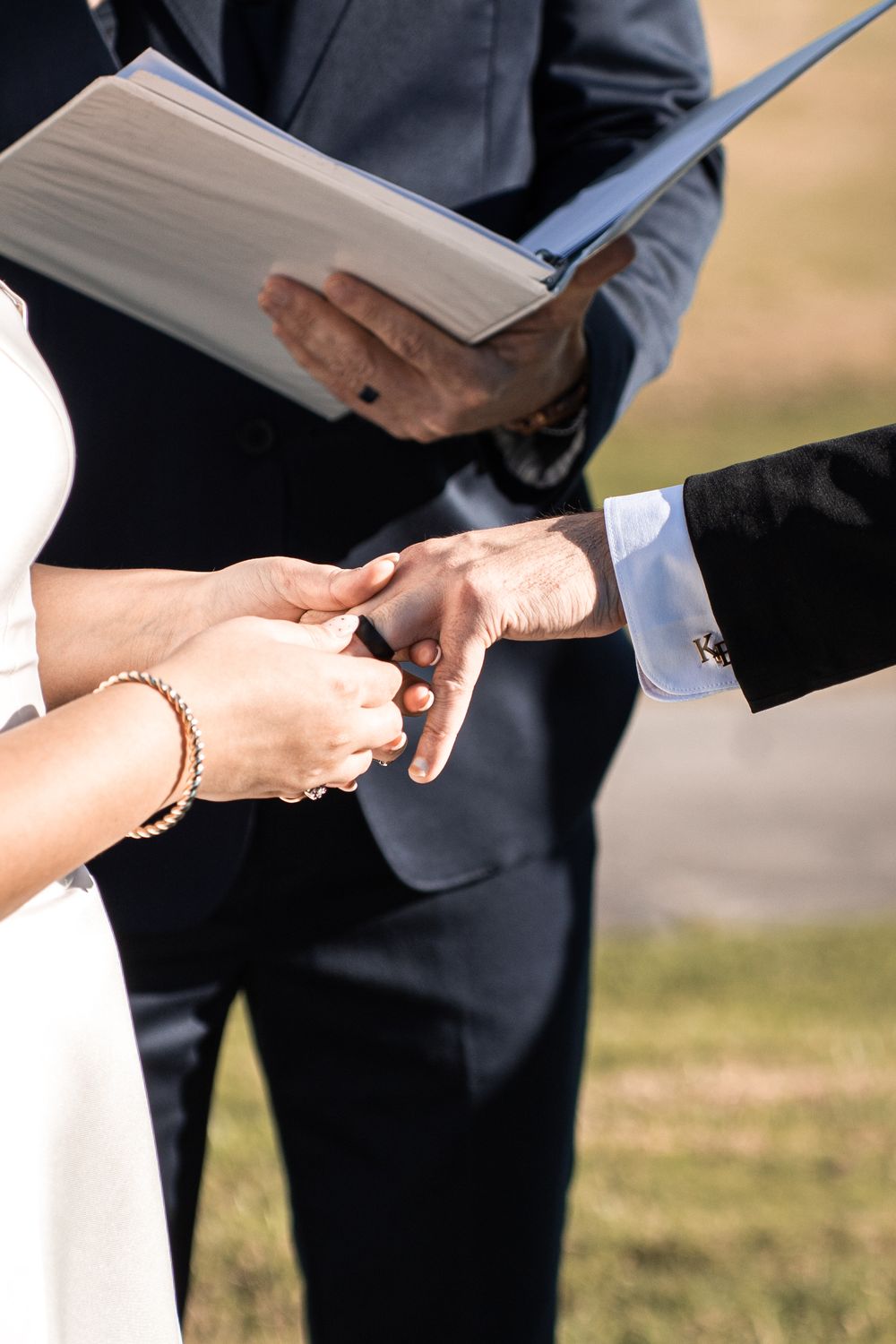 Bride and groom exchanging rings during outdoor wedding ceremony while officiant holds ceremony book.
