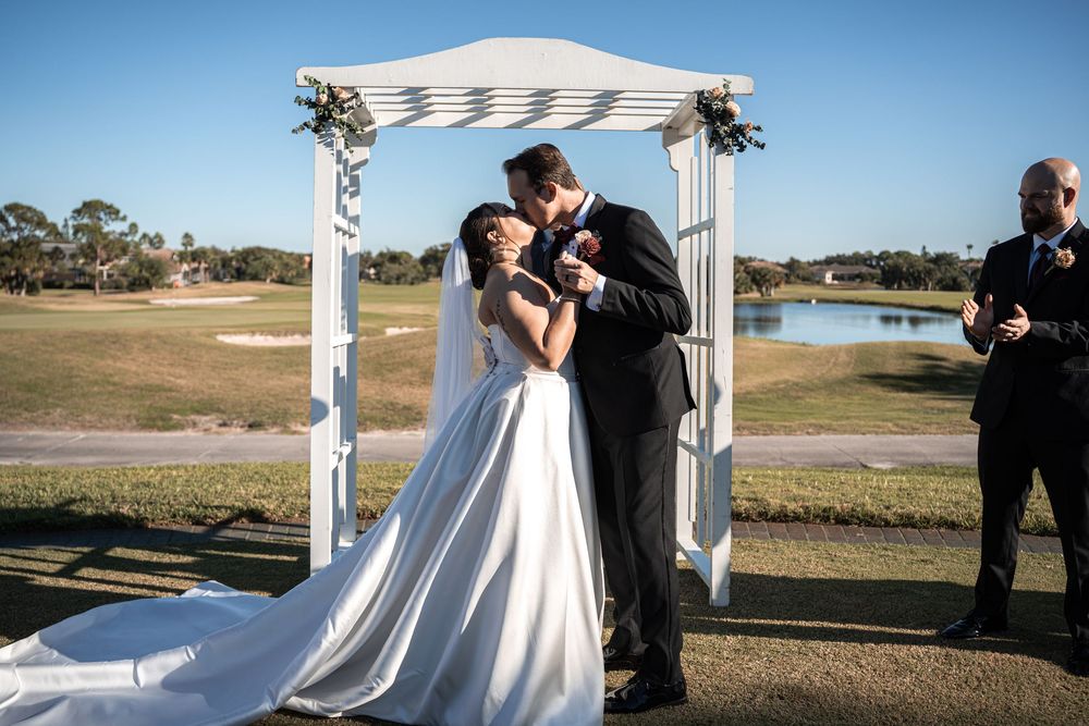 Bride and groom share first kiss under white floral arch at outdoor golf course wedding ceremony.
