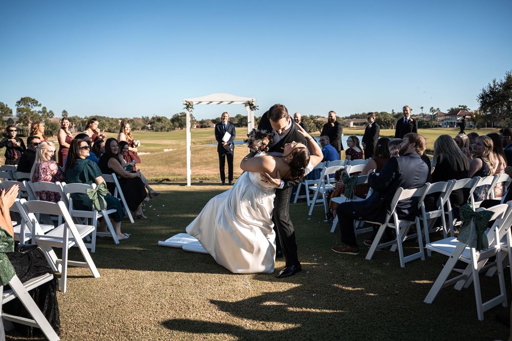 Bride and groom share a romantic dip kiss at outdoor wedding ceremony with white chairs and arch on sunny day.