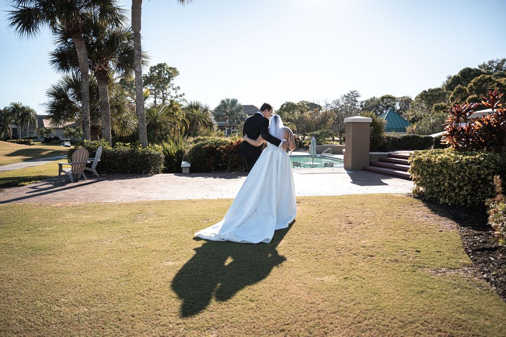 Bride and groom dancing on green lawn near pool with palm trees at outdoor wedding venue.