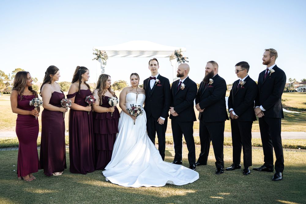 Wedding party poses outdoors, bridesmaids in burgundy gowns and groomsmen in black suits flanking bride and groom.