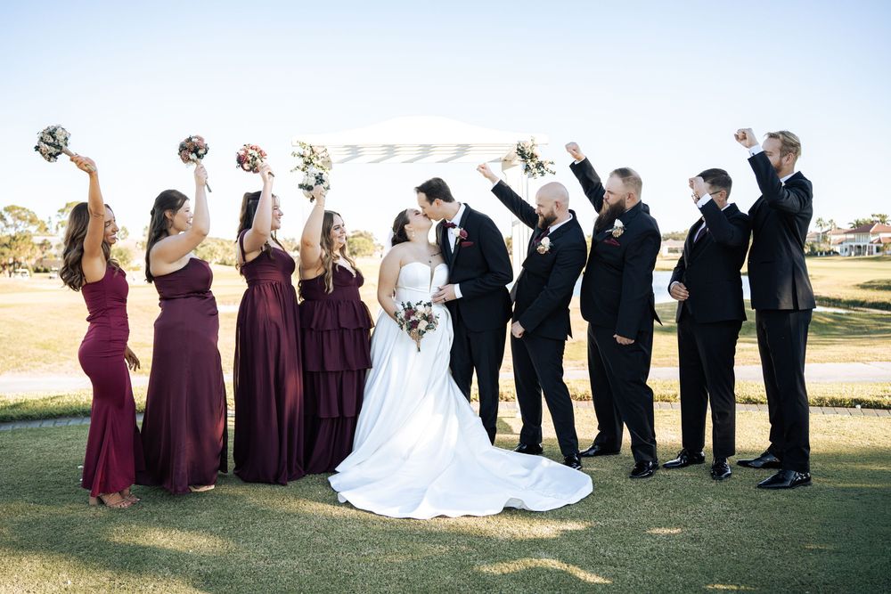 Wedding party celebrates as bride and groom kiss under a white arch on a golf course, bridesmaids in burgundy gowns.