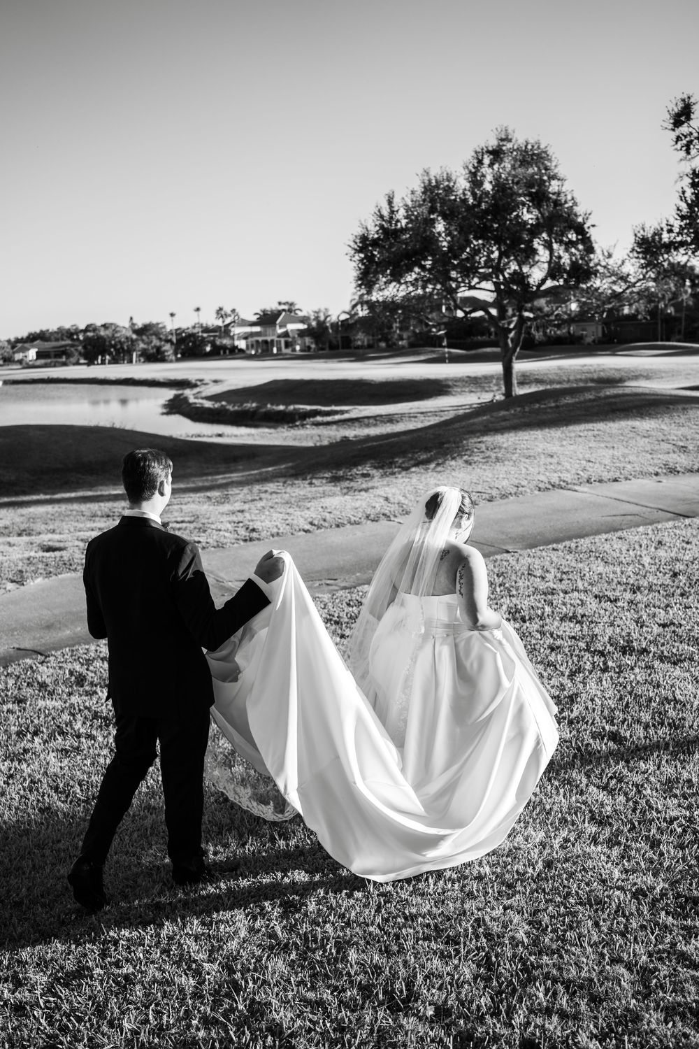 Black and white photo of bride and groom walking hand in hand across a golf course, bride's veil flowing behind.
