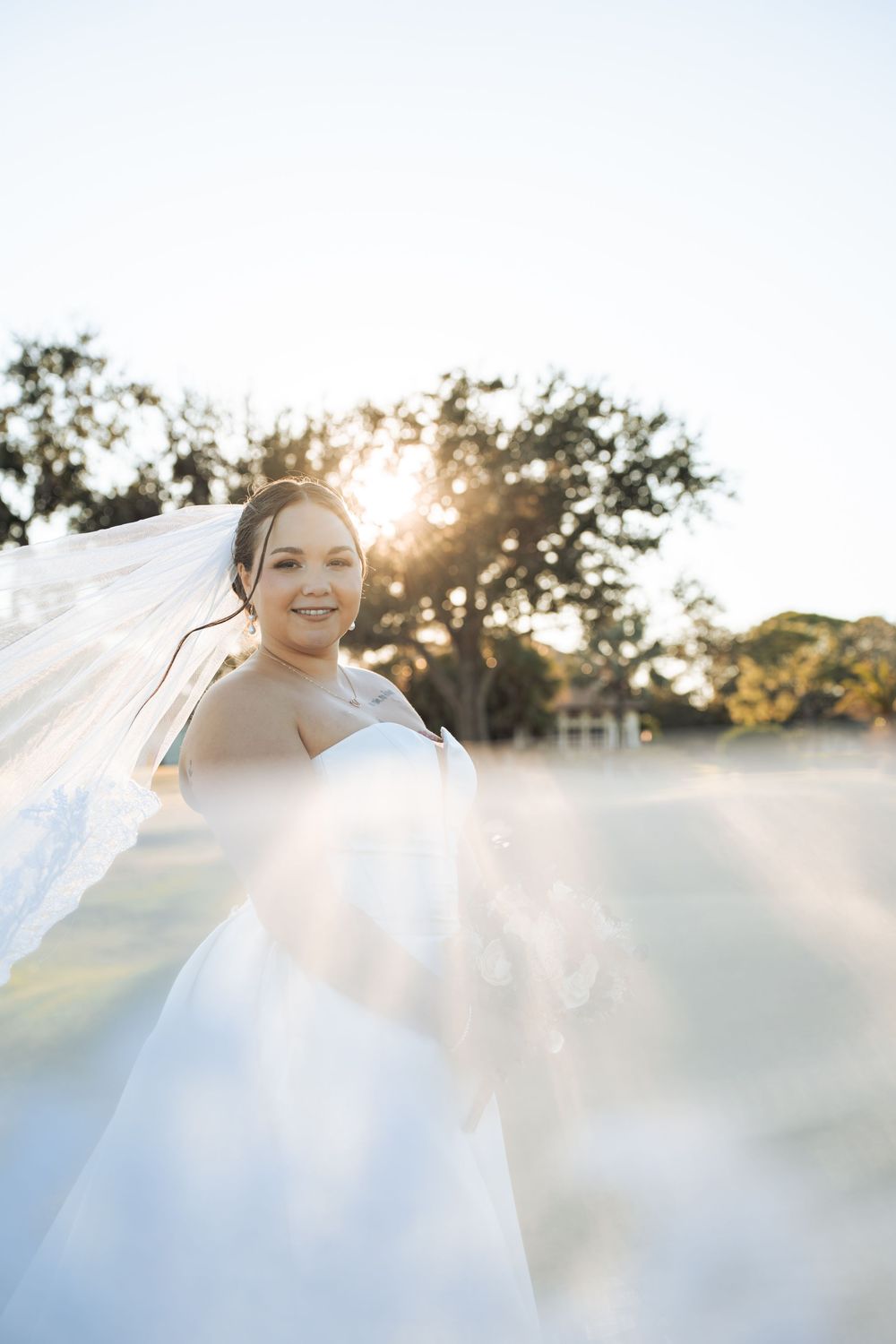 Bride in white gown and flowing veil smiling outdoors with golden sunlight and trees in the background.