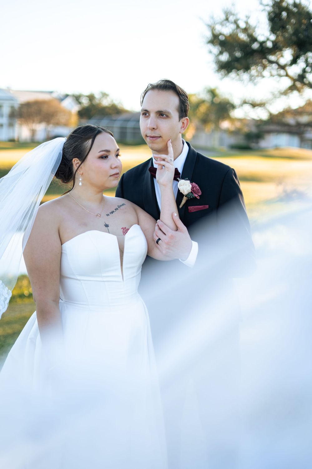 Bride in strapless white gown and groom in black tuxedo pose together at outdoor wedding.