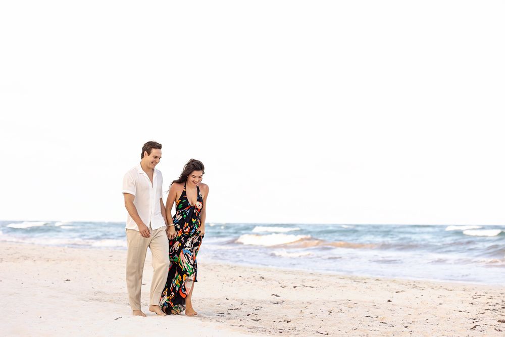 Couple walking hand in hand on a sandy beach, woman in floral maxi dress, man in white shirt and tan pants.