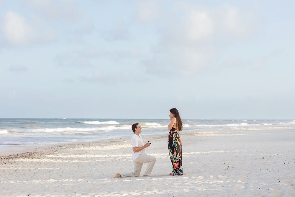 Man on one knee proposing to woman on a serene white sand beach with ocean waves behind them at The Beach Tulum Hotel.