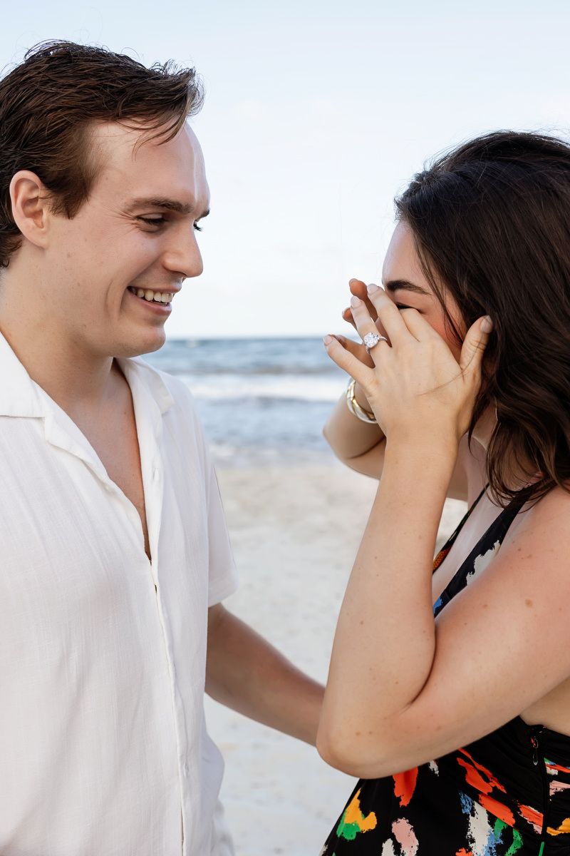 Joyful couple sharing an engagement moment on the beach, woman showing off her new ring while smiling.