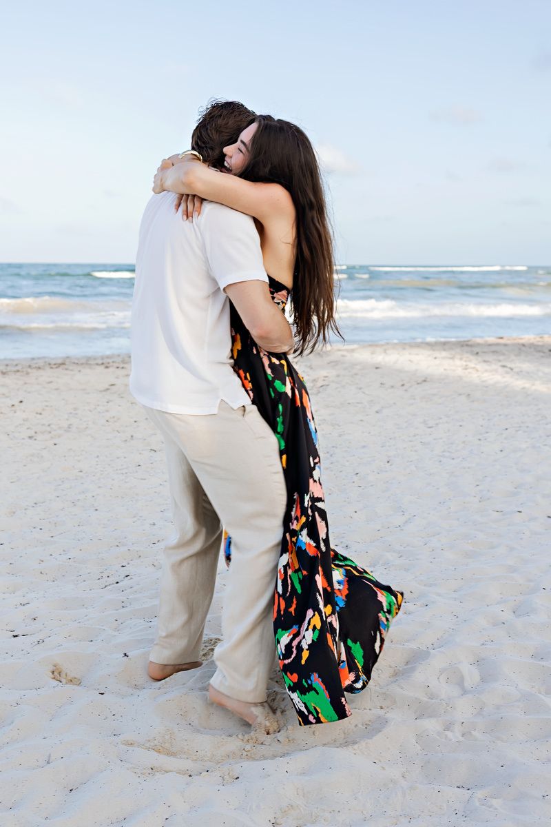 Couple embracing romantically on a sandy beach with ocean waves in the background, woman in colorful maxi dress.
