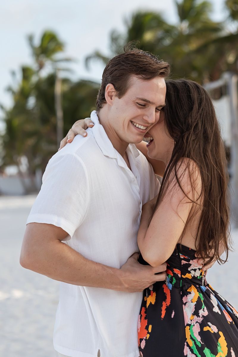 Happy couple embracing and smiling on a tropical beach, woman in colorful dress, man in white shirt, palm trees background.