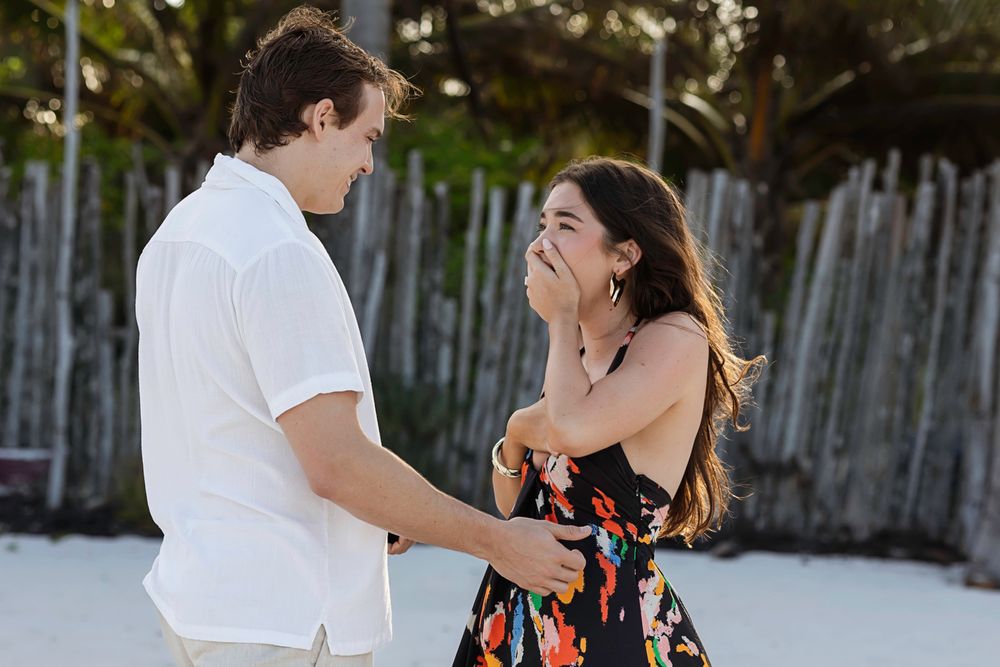Surprised woman covers mouth as man proposes on a sunny tropical beach.