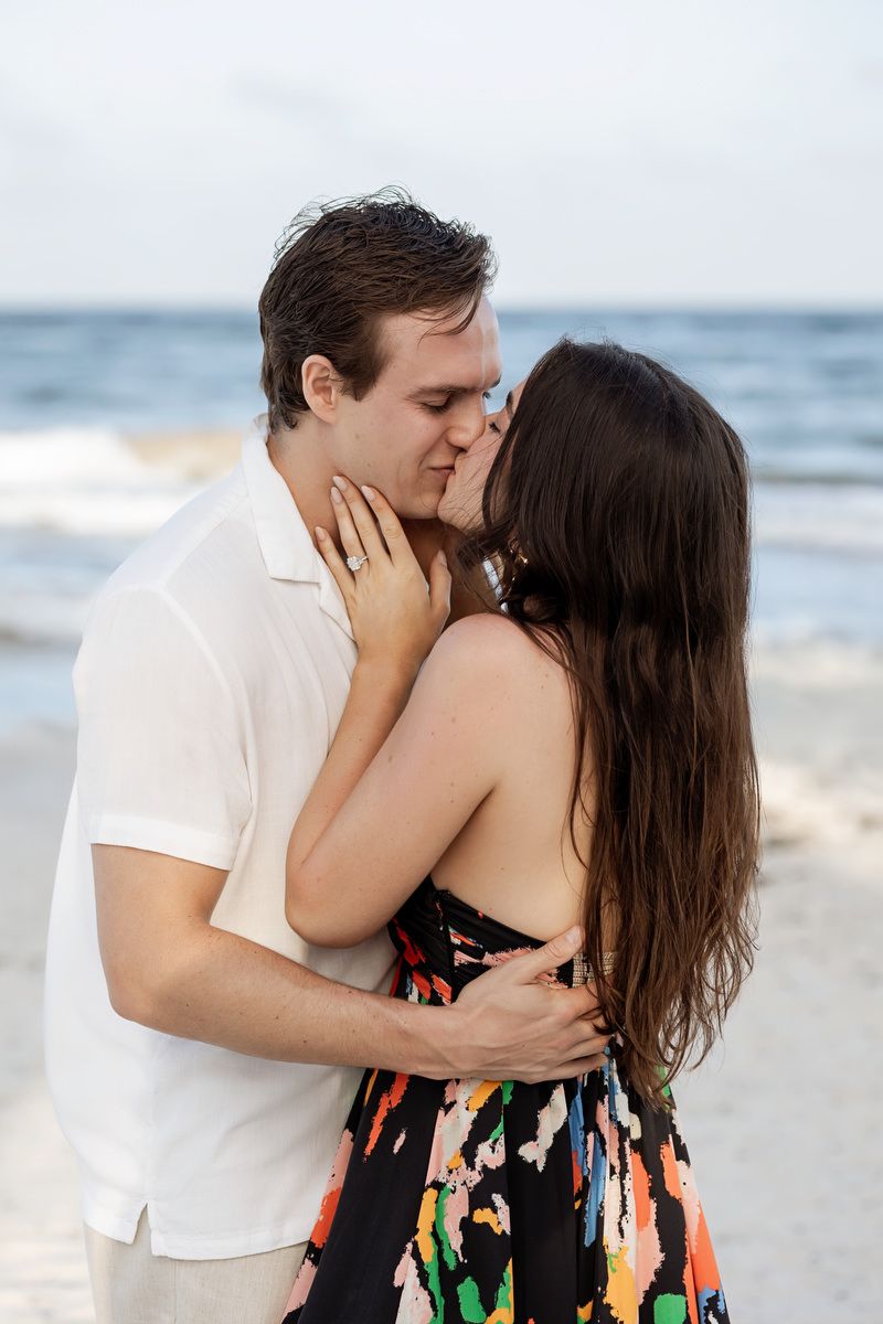Couple sharing a romantic kiss on a beach, woman in colorful floral dress showing engagement ring.