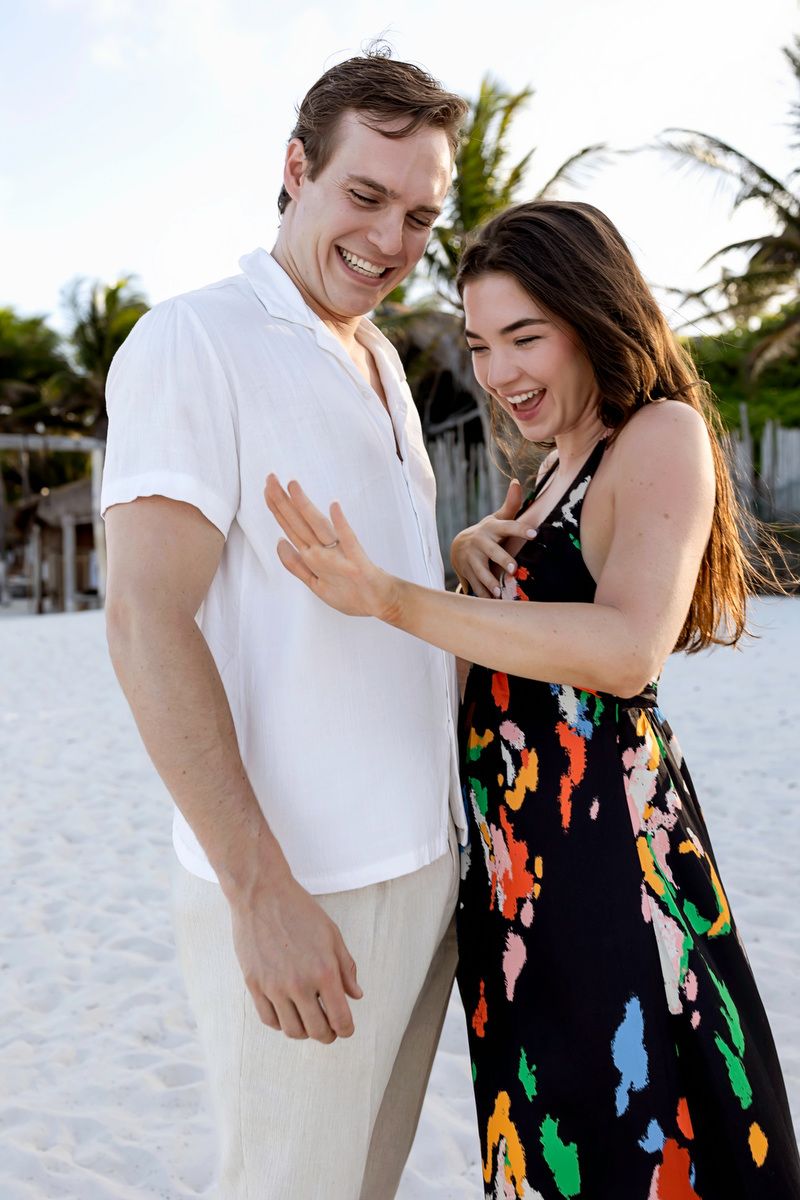 Joyful couple laughing together on white sand beach, woman in colorful dress showing engagement ring.