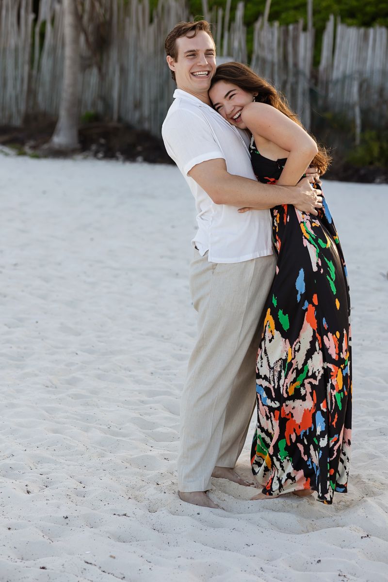 Happy couple embracing on a white sand beach, woman in colorful maxi dress hugging man in white shirt and khaki pants.