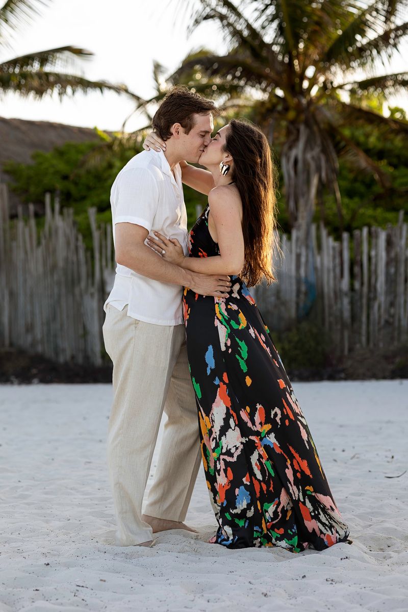 Happy couple sharing romantic kiss on sandy beach with palm tree backdrop at sunset.