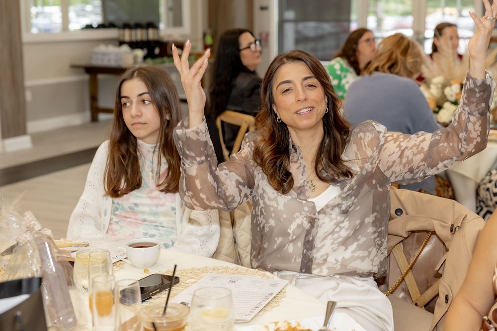 Two women sitting at a table at an indoor event, one raising her hand while smiling.