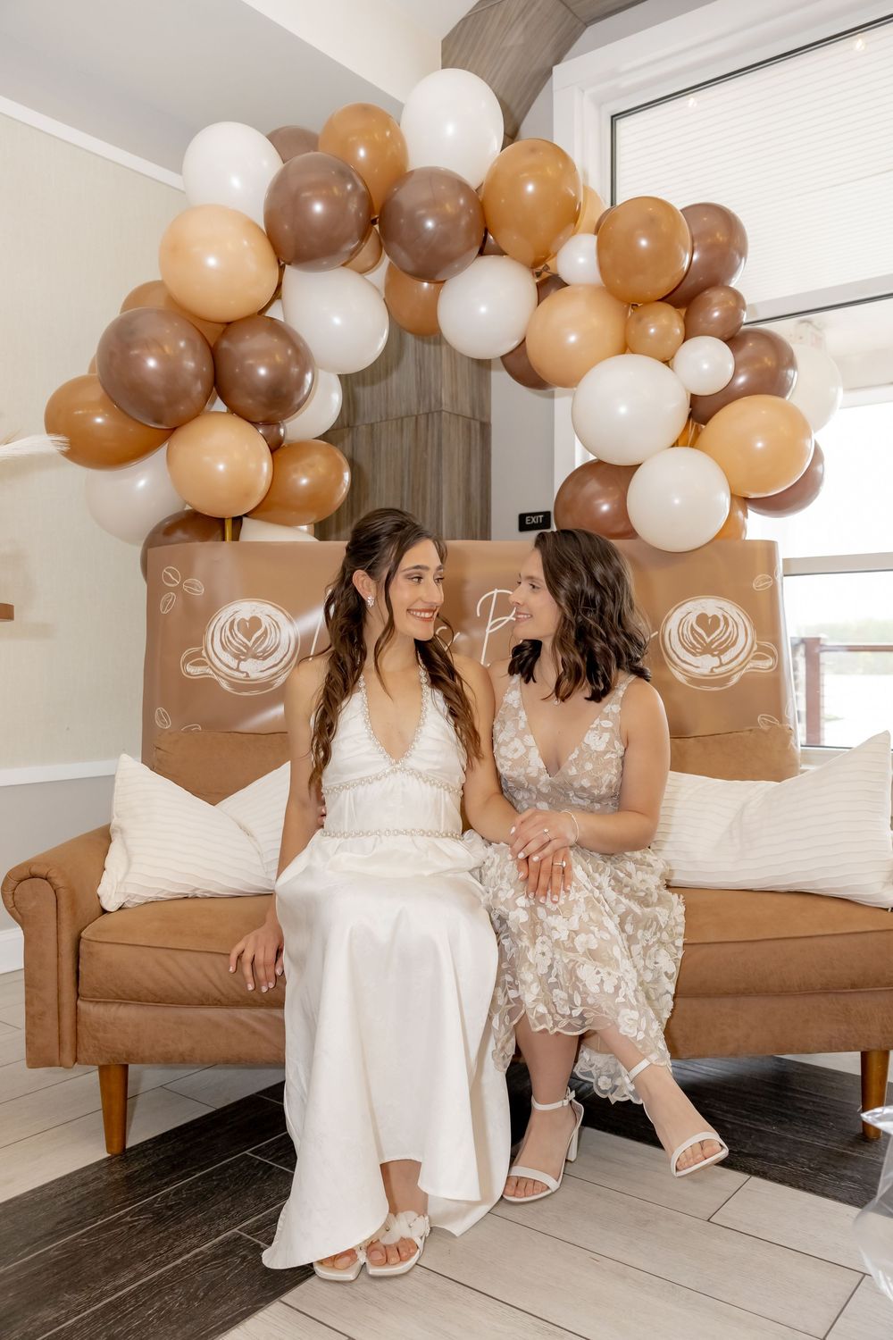Two brides sit on a tan sofa under a brown, white, and beige balloon arch at their bridal shower celebration.