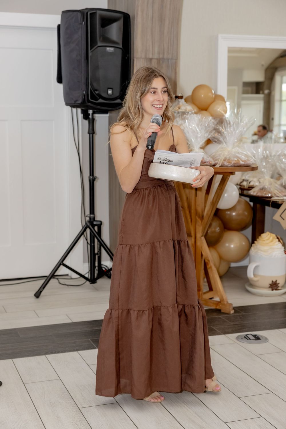 Woman in brown maxi dress speaking into microphone at indoor event with balloon decorations and speaker stand.