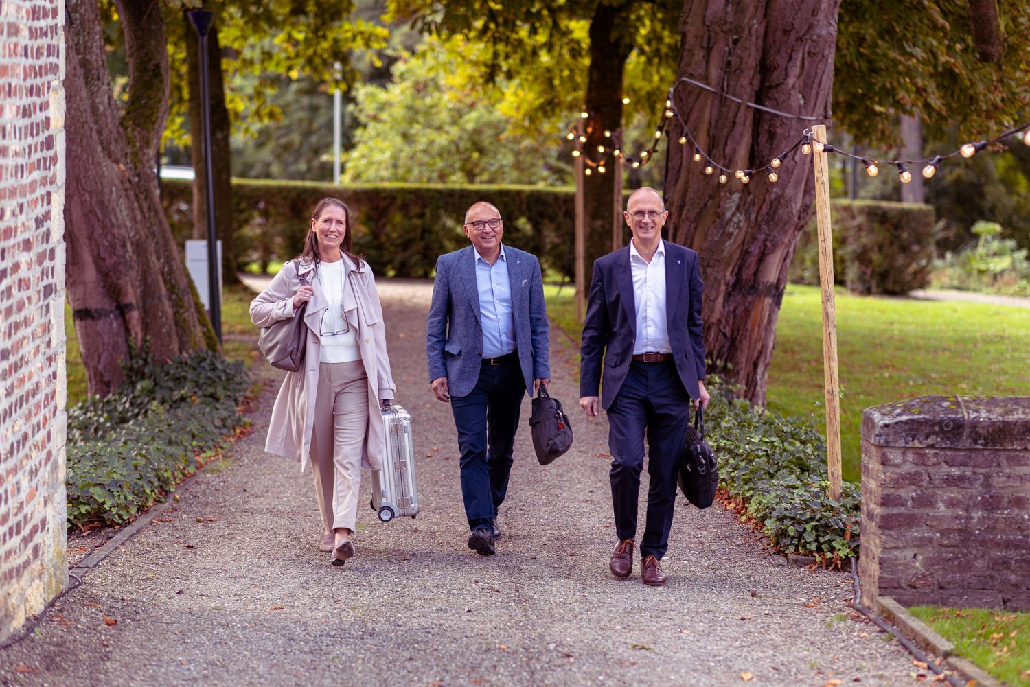 Three business professionals walking confidently on a garden path surrounded by tall trees and string lights.
