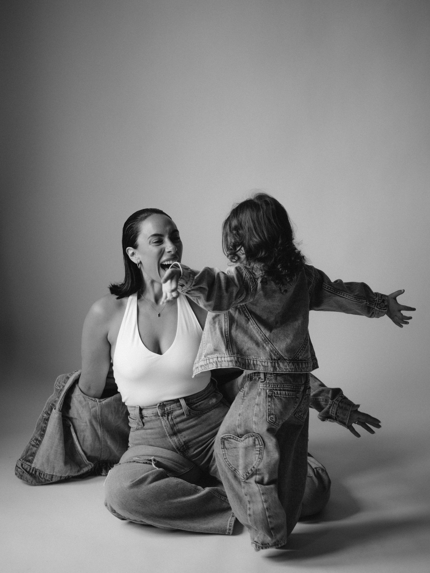 Black and white photo of two women in denim sitting on floor, one laughing as the other gestures expressively.