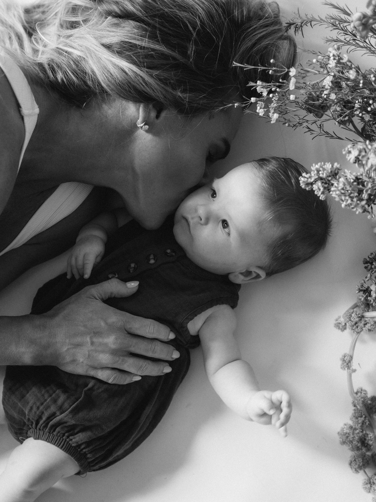Black and white photo of a woman kissing a newborn baby lying among delicate wildflowers.
