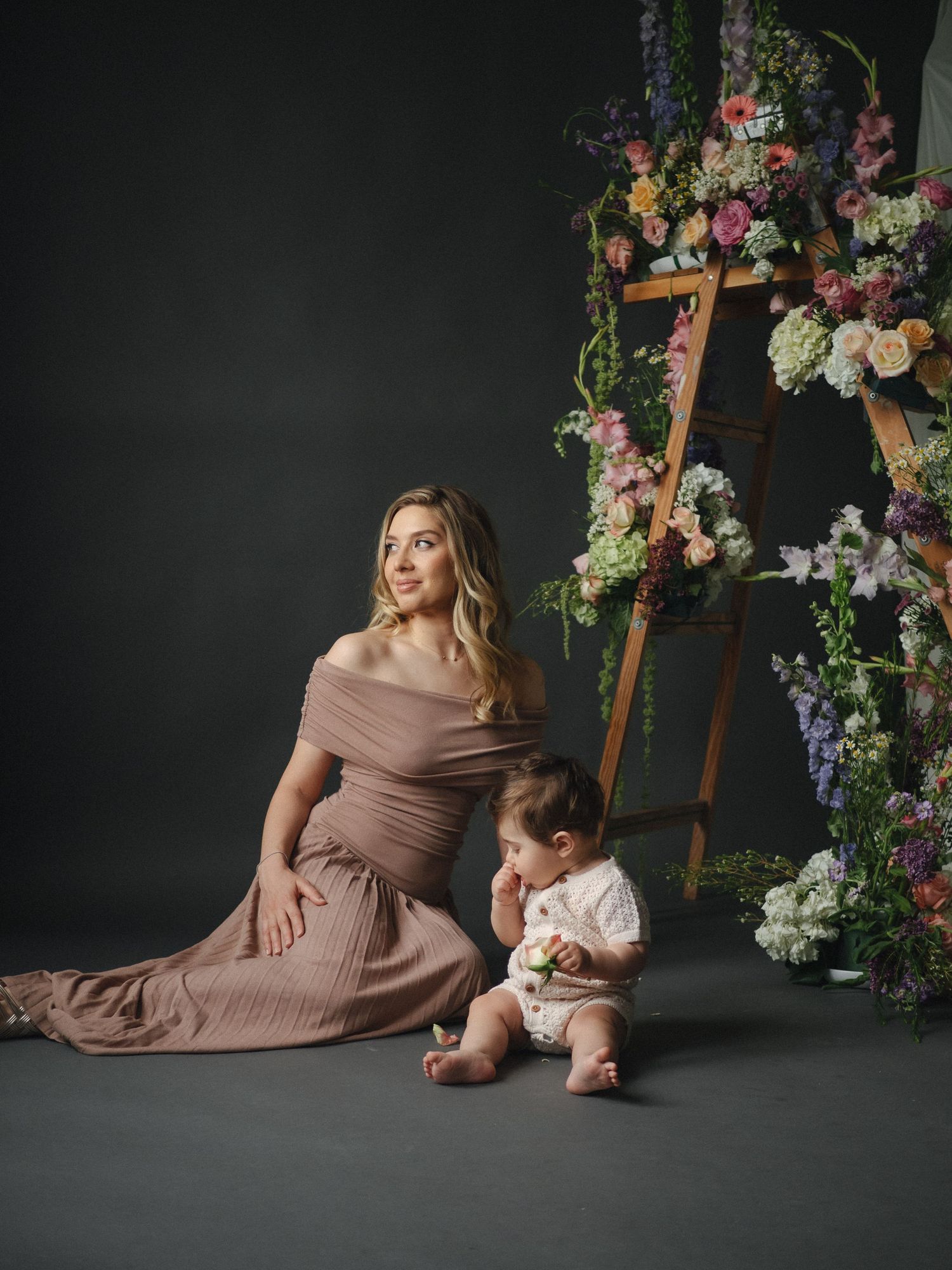 A woman in a mauve off-shoulder gown sits beside a baby near a floral-adorned wooden ladder on a dark studio backdrop.