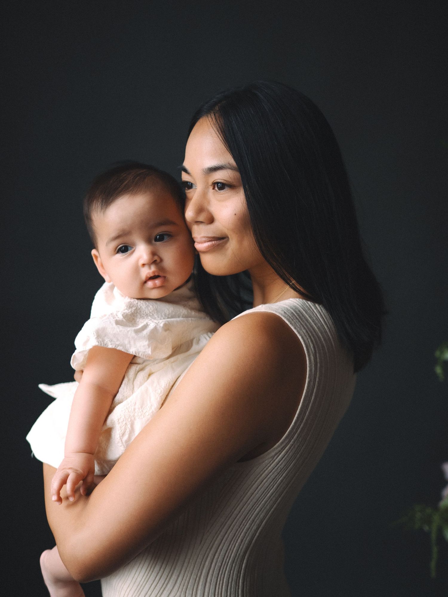 Asian mother in white dress holds infant baby against dark background in tender portrait photo.