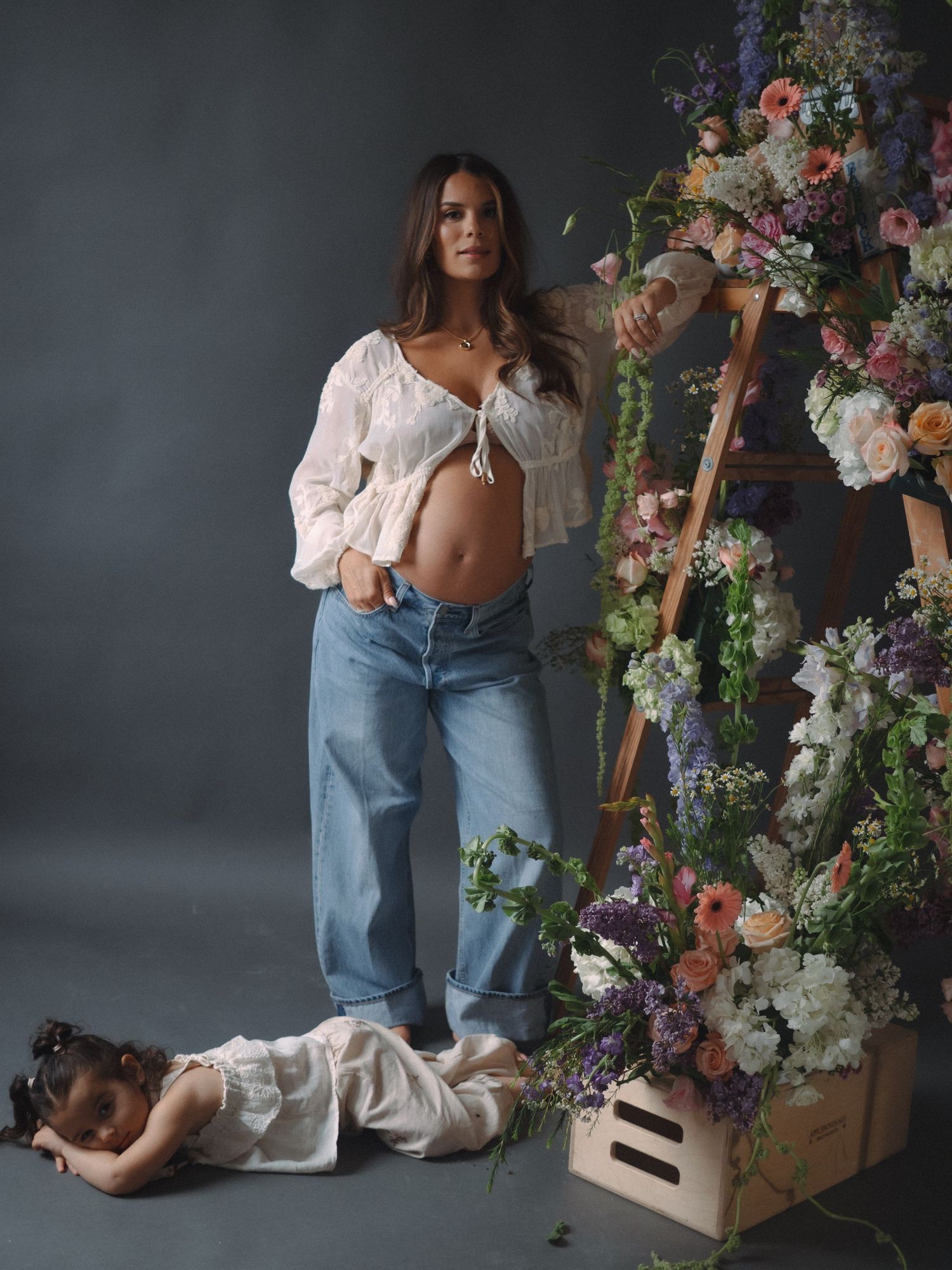Pregnant woman in white top and jeans poses near floral ladder display with child lying on studio floor.