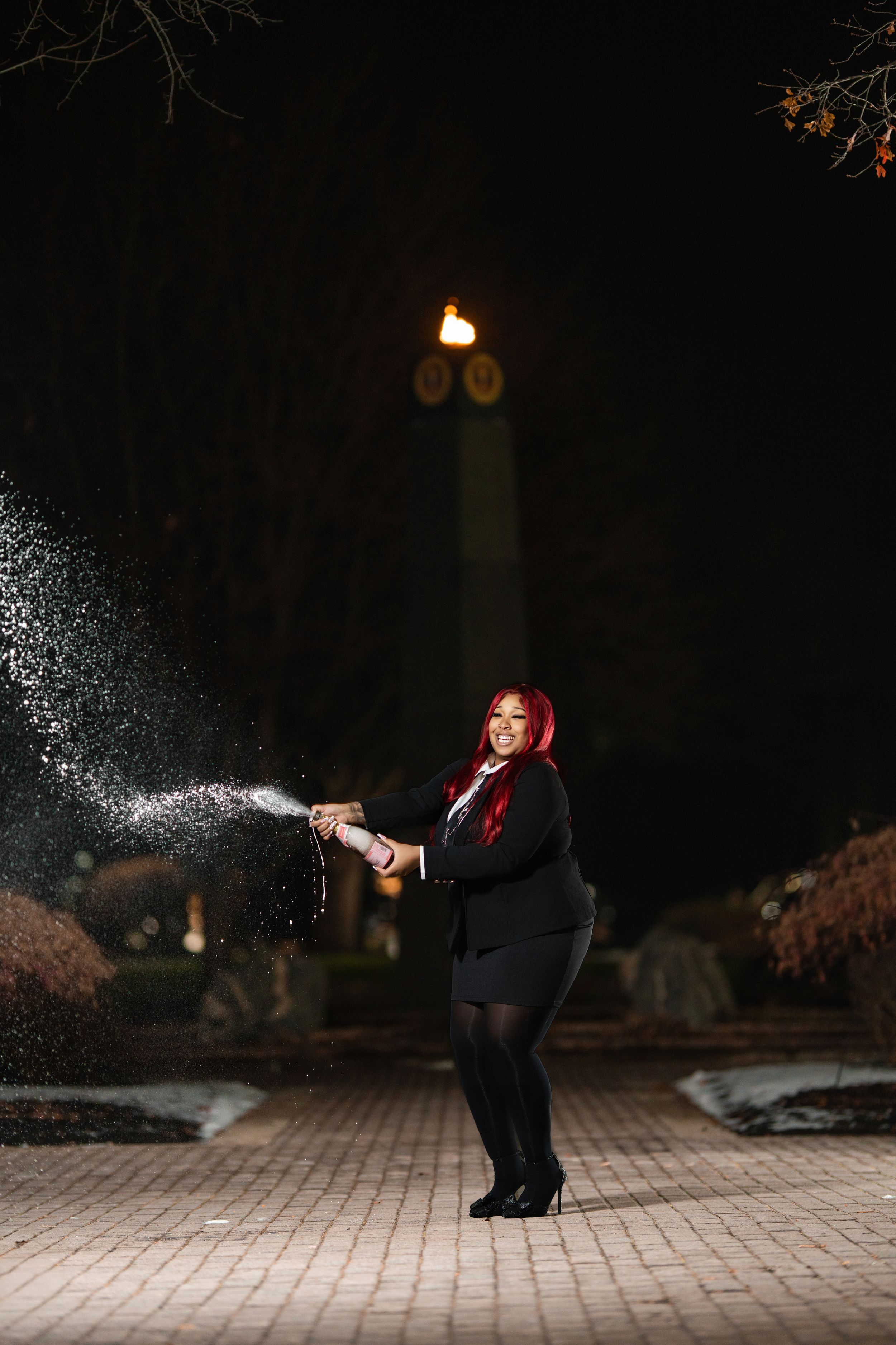 Woman with red hair in black suit sprays champagne bottle outdoors at night near lit monument.