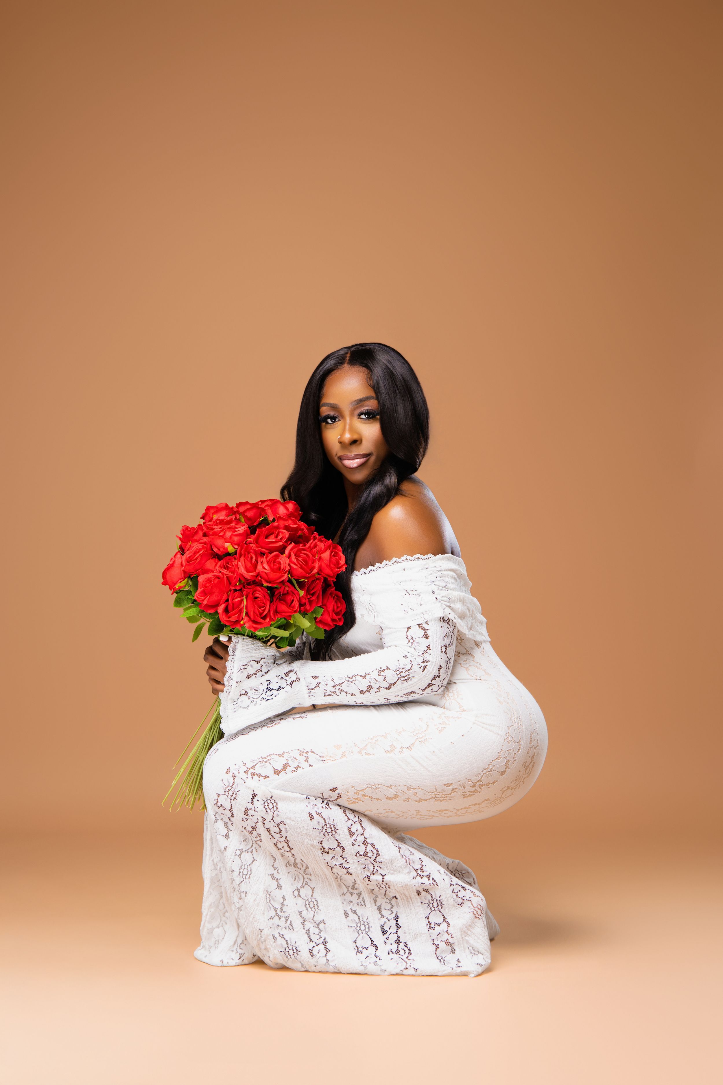 Smiling woman in white lace dress crouches while holding large bouquet of red roses on beige background.
