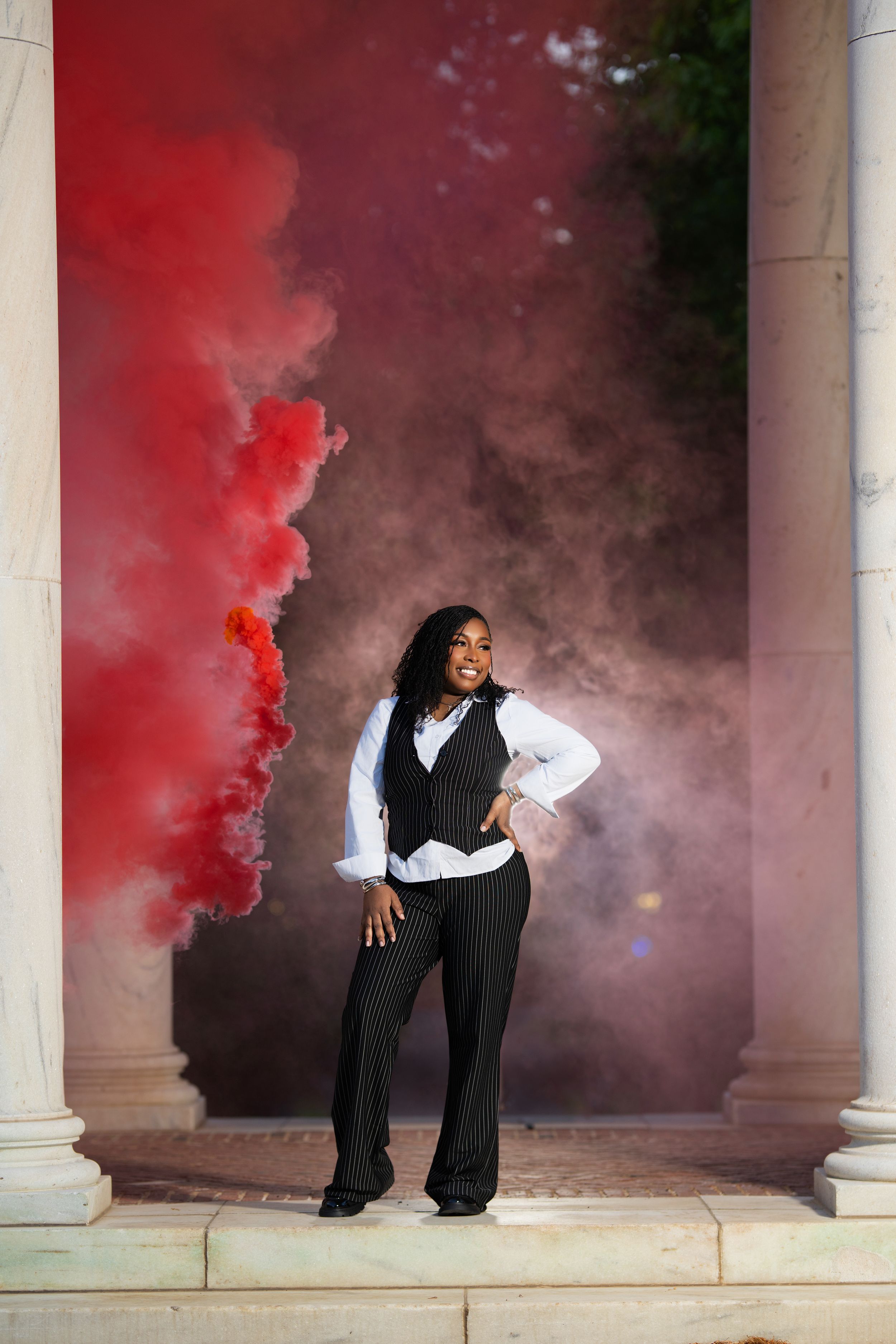 Woman in vest and slacks poses amid red smoke between white columns.