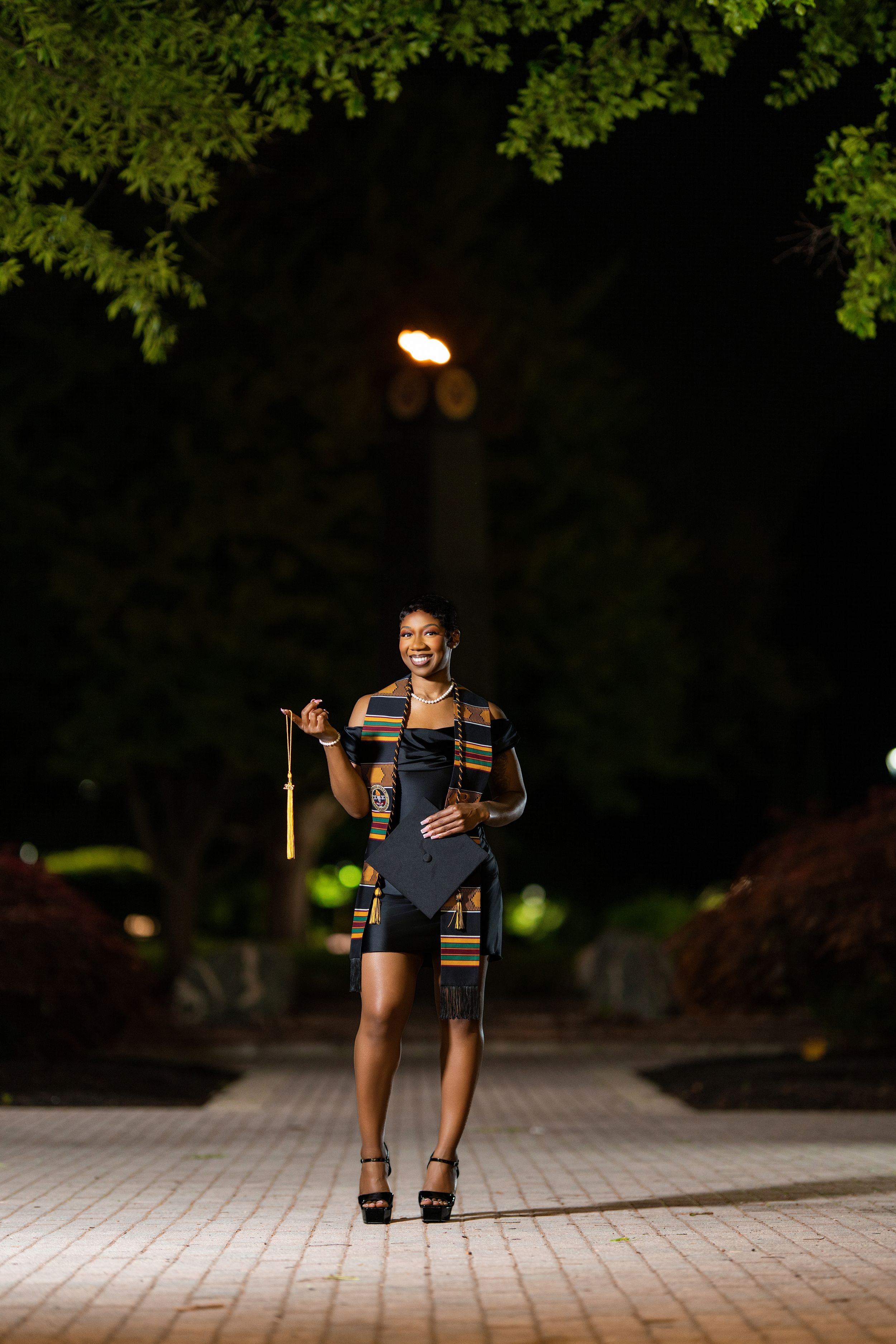 Joyful graduate in kente stole holds cap while walking at night.