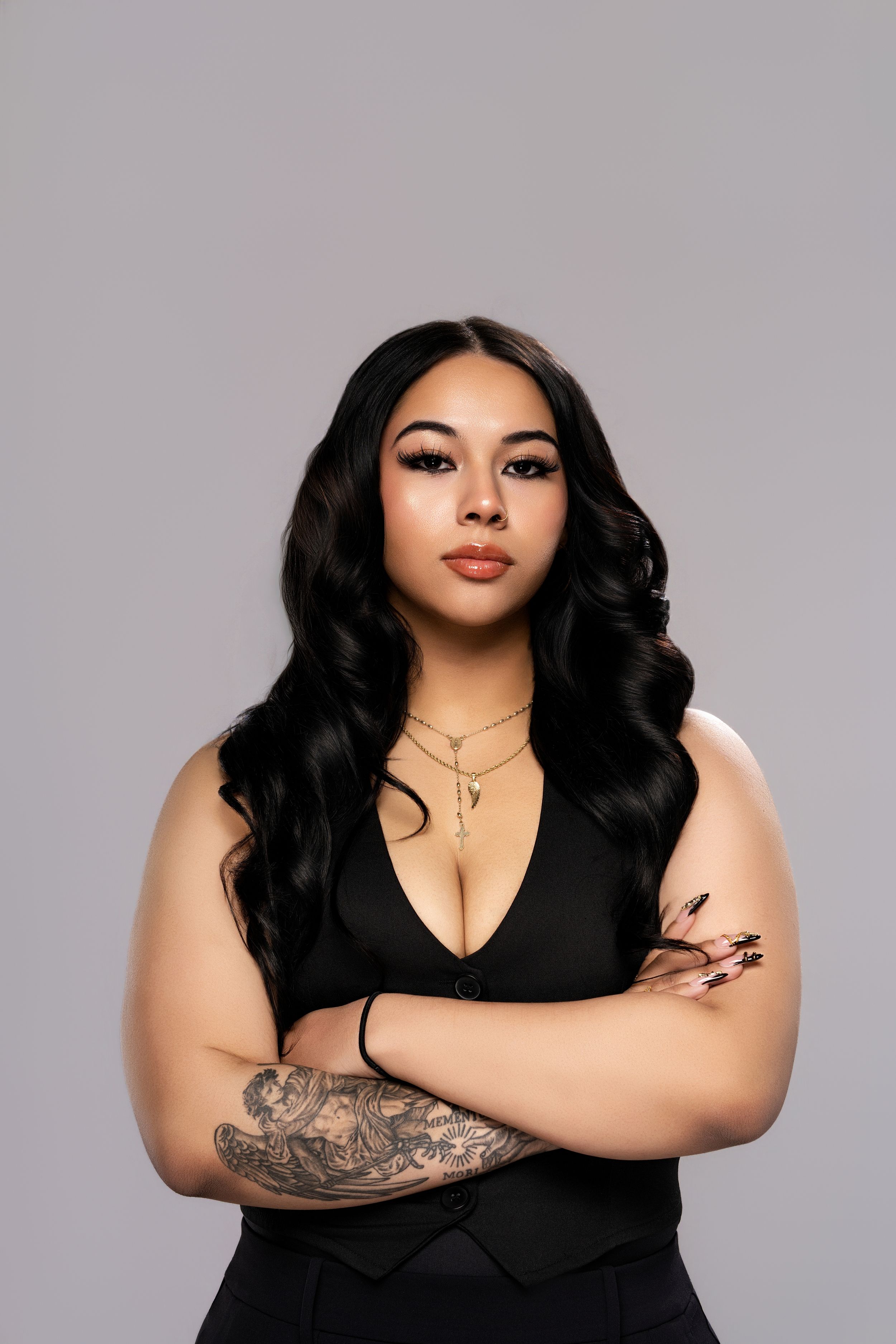 Woman in black dress with tattoo and gold necklaces poses confidently with arms crossed against gray background.