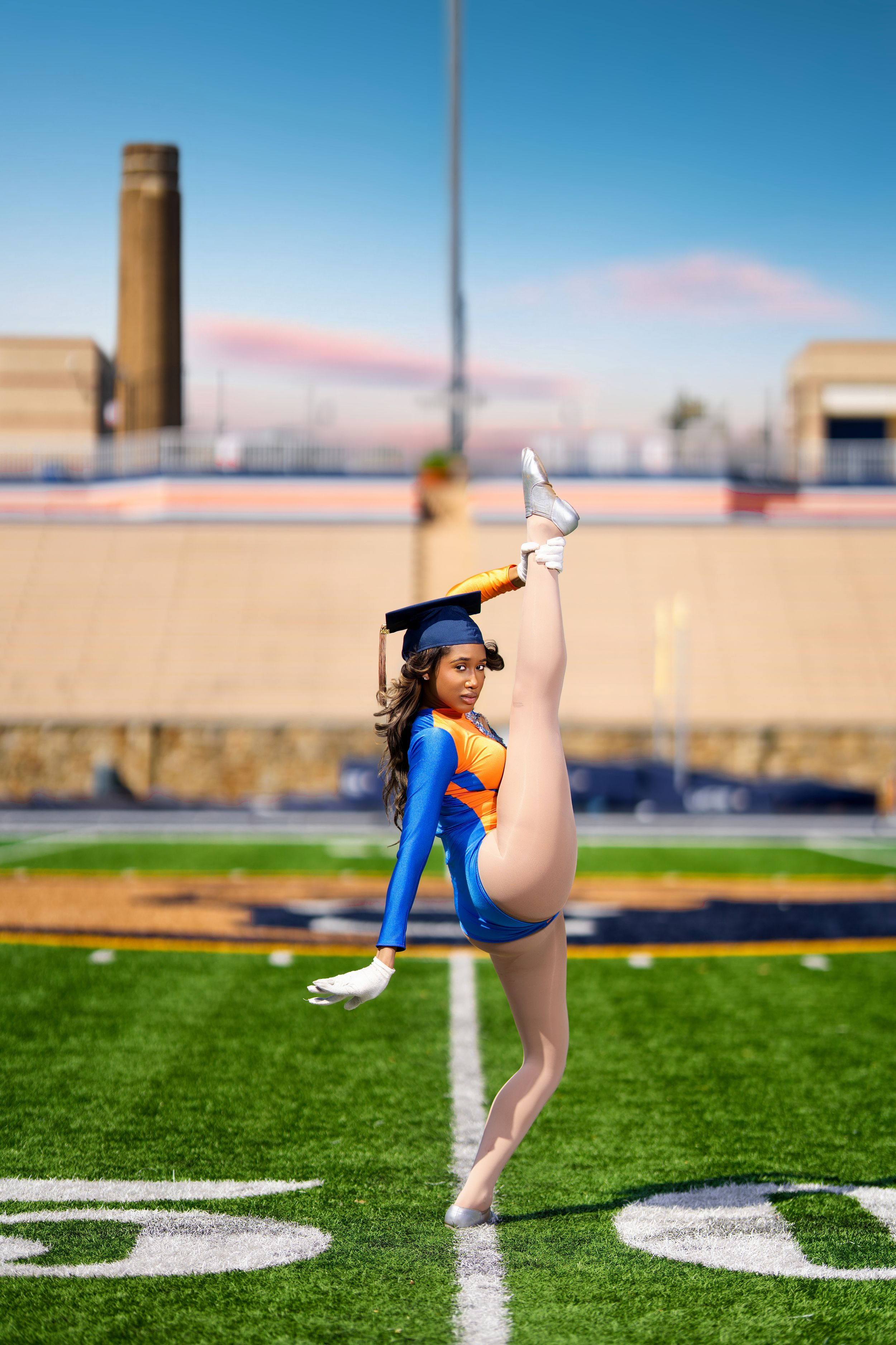 Flexible dancer in graduation cap performs high kick on football field.