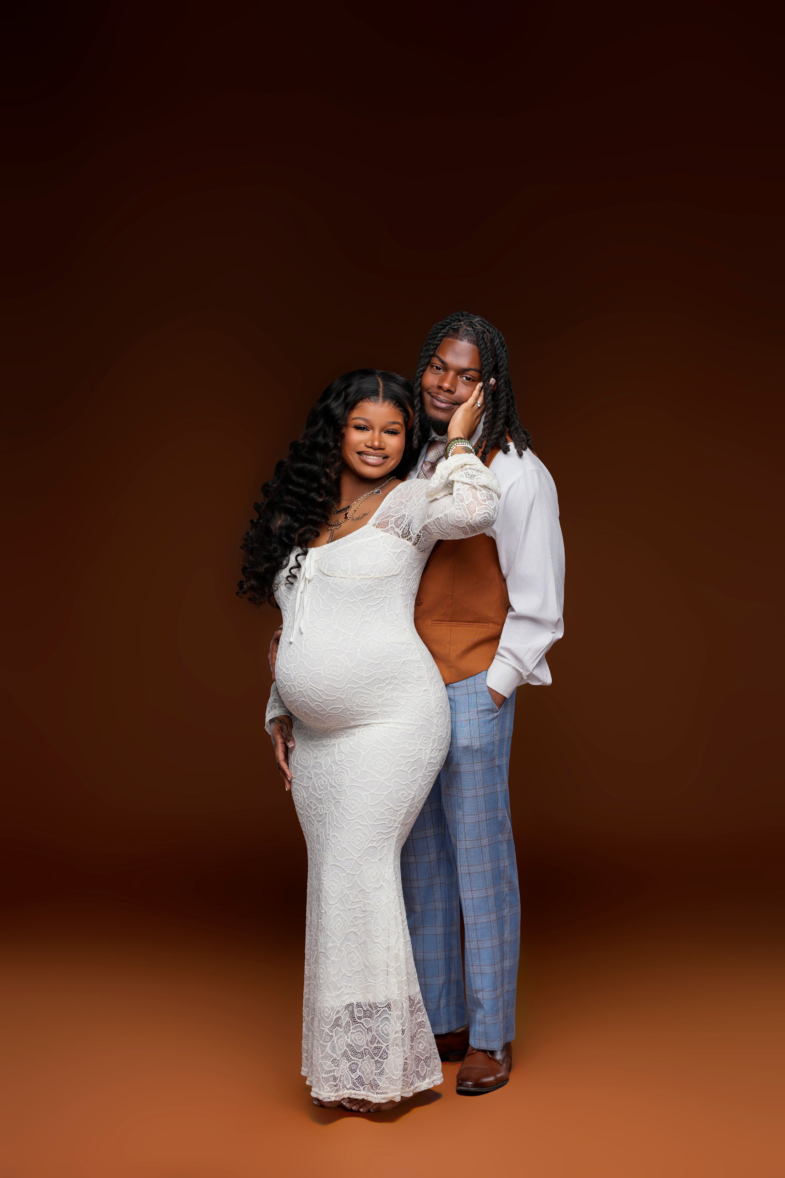 Expecting couple poses together, woman in white lace maternity gown, man in white shirt, brown background.