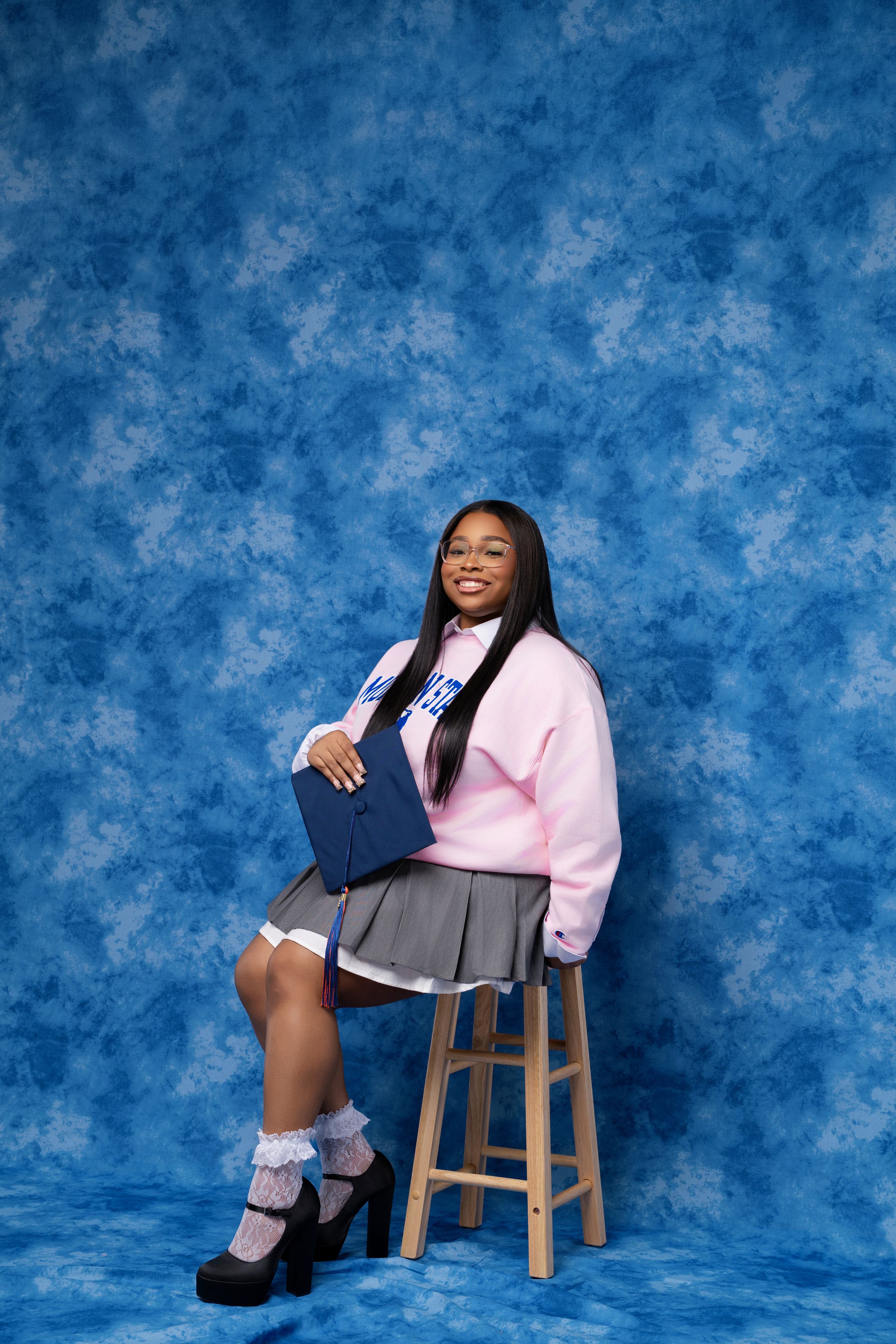 Young woman in pink sweatshirt and plaid skirt poses on stool with blue backdrop.