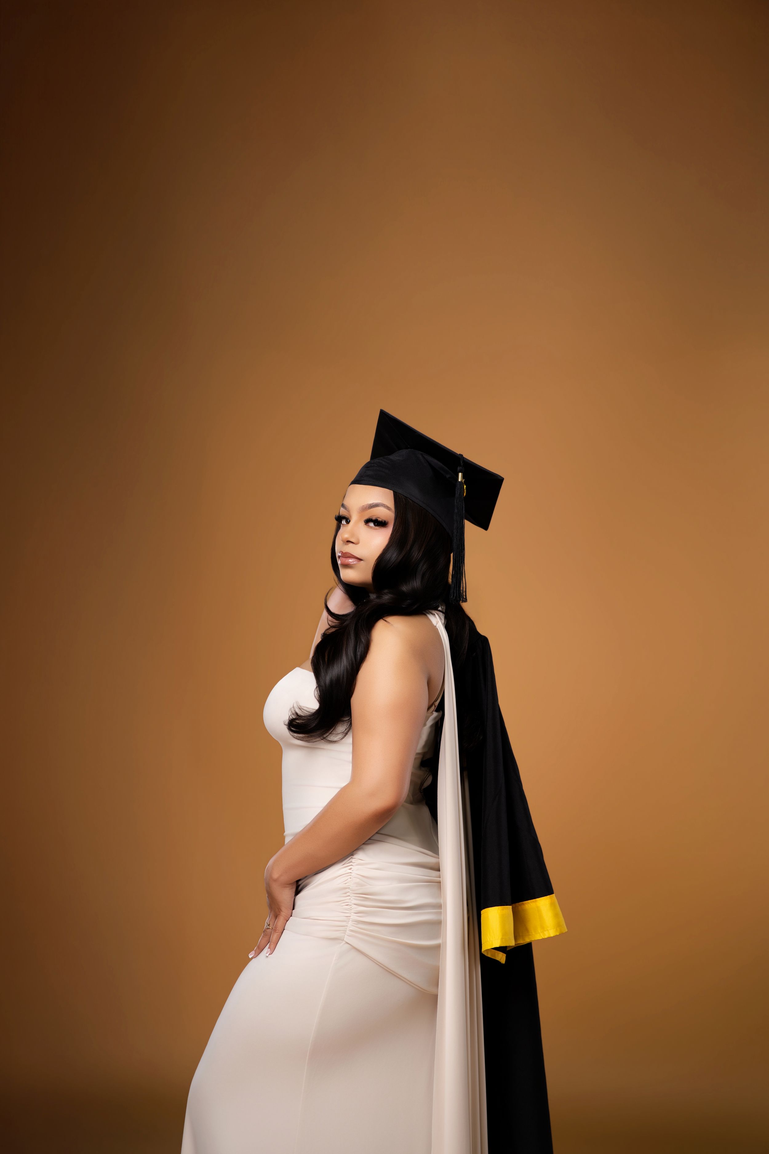 Graduate in white dress and black cap with gold trim poses elegantly against warm golden brown background.