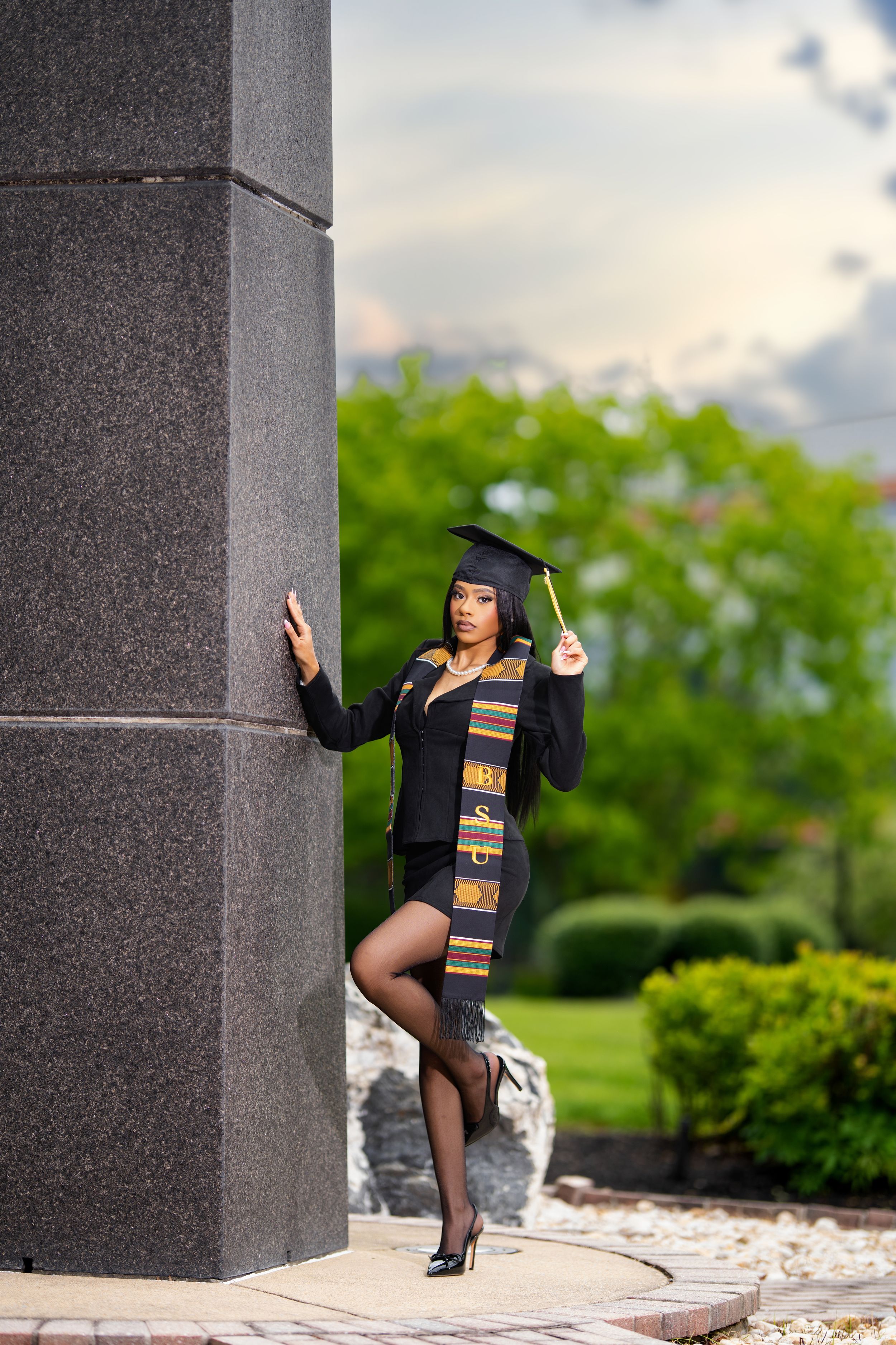 Female graduate in cap and gown leans against stone pillar outdoors.